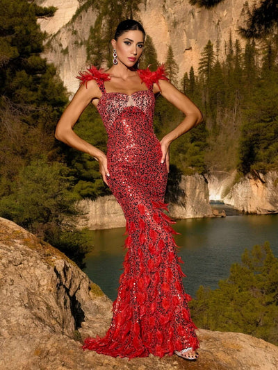 Woman wearing Red Glitter Sequins Maxi Dress with feather details standing in front of a scenic rocky landscape and lake.